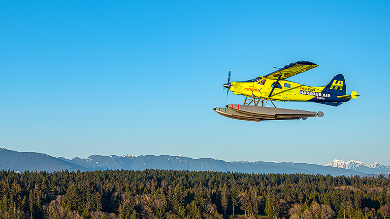 A yellow seaplane with "Harbour Air" branding is flying above a forested area, with a background of distant mountains under a clear blue sky.