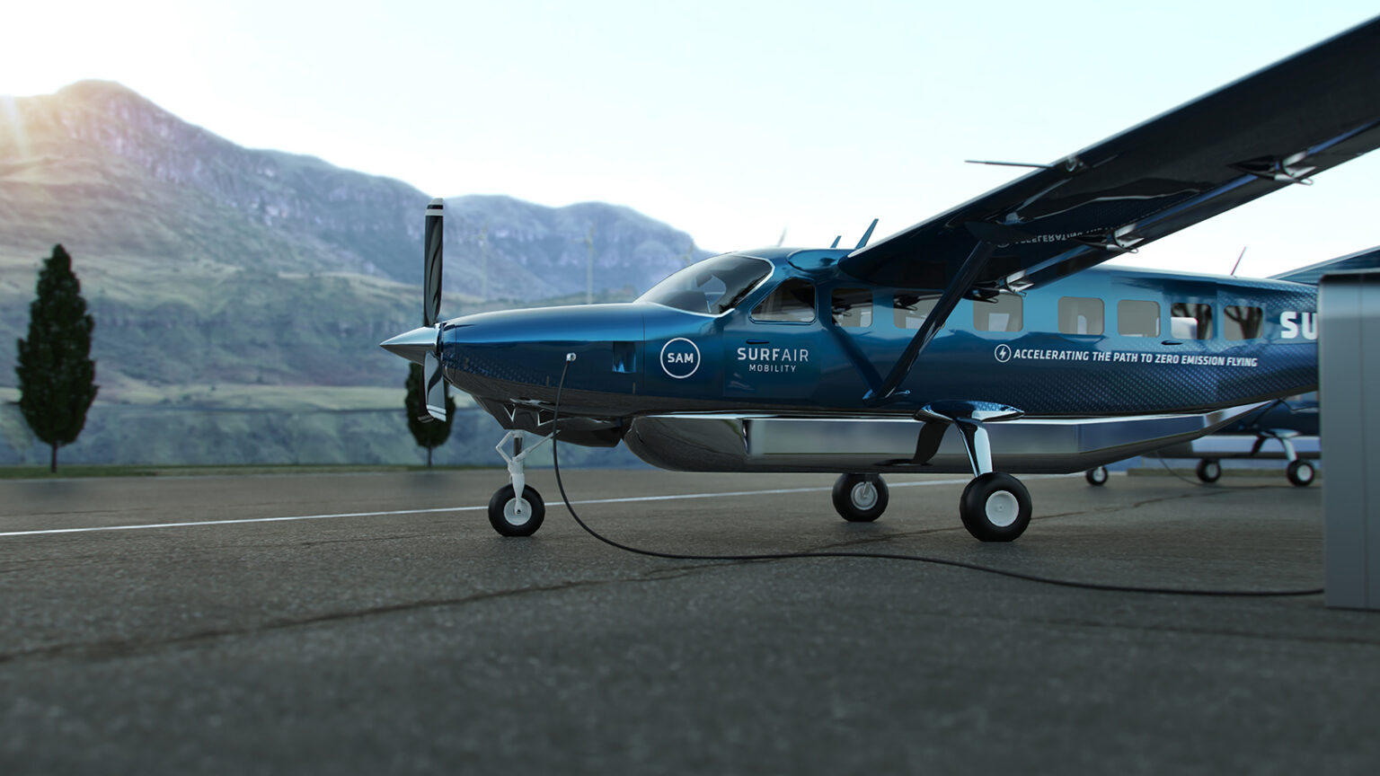 A blue single-engine turboprop airplane is parked on a tarmac against a backdrop of mountains and trees. The aircraft is connected to ground power.