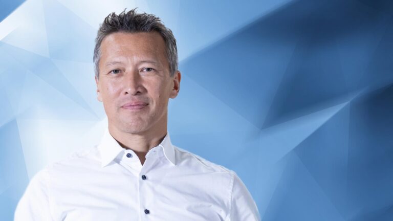A person with short gray hair wearing a white button-up shirt stands against a blue geometric background.