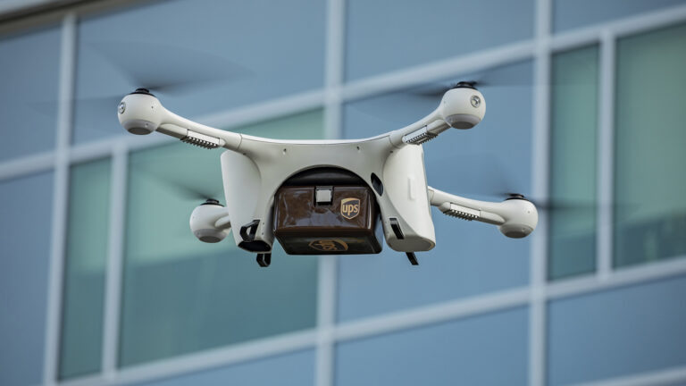 A white drone with four propellers is carrying a small brown UPS package as it flies in front of a windowed building.