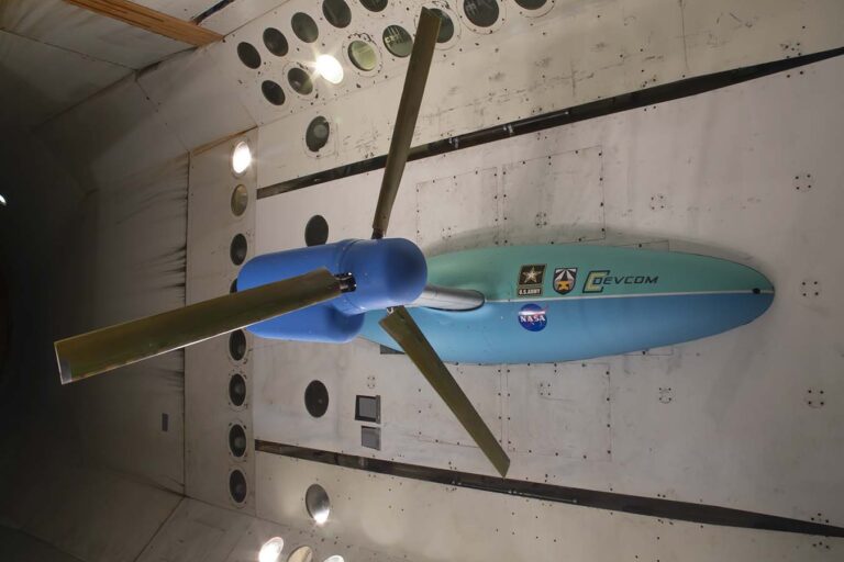 A large teal and blue rotorblade model is suspended in a wind tunnel for testing. Various organization logos, including NASA, are displayed on the model.