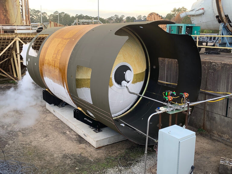 A cylindrical industrial container with attached pipes and valves is undergoing a pressure test, emitting steam or gas. The background includes industrial equipment and structures.