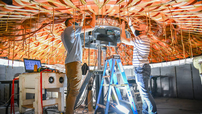 Two people work on wiring inside a dome structure, with one person on a blue ladder and the other standing. Equipment and tools are visible around them.