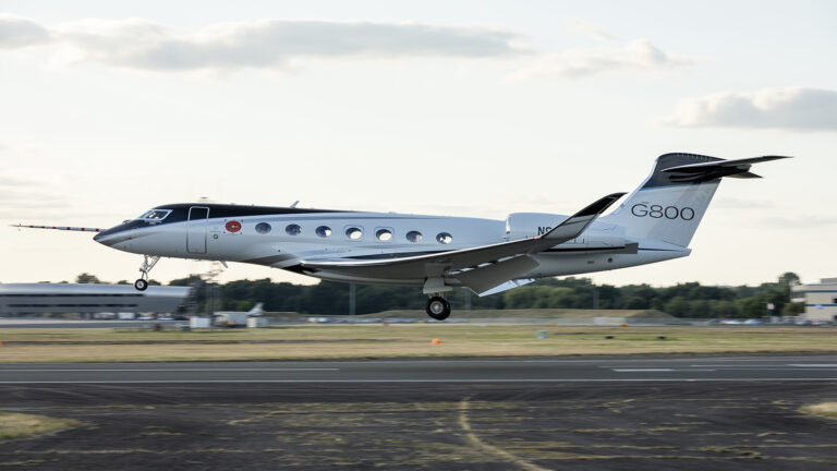 A Gulfstream G800 aircraft taking off from a runway, with its landing gear extended and several buildings visible in the background.