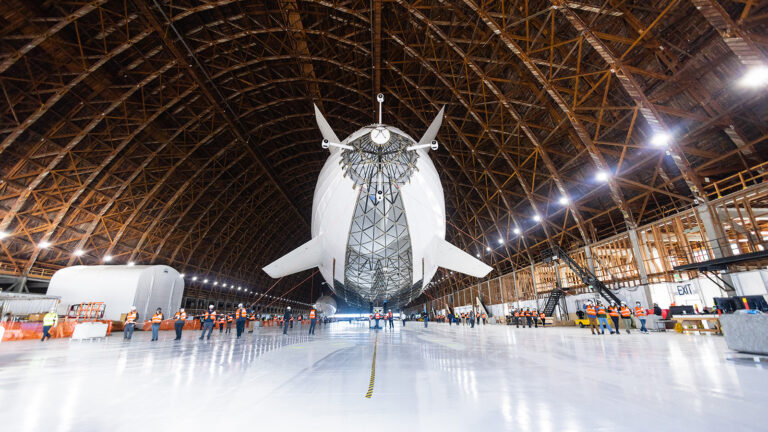 A large airship is stationed inside a spacious hangar with a high, arched wooden ceiling. Several people in safety vests and helmets are gathered around it, and equipment is scattered throughout.