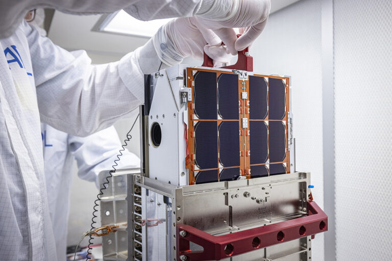 Technicians in white attire handling a small satellite with solar panels inside a cleanroom environment.