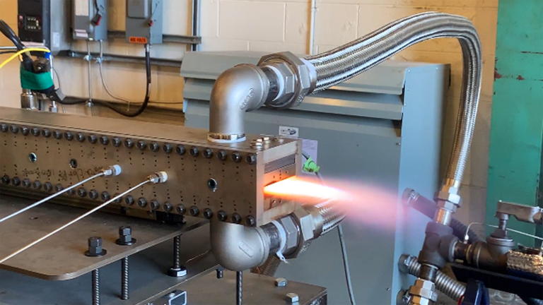 A test setup with a metal apparatus emitting a controlled flame or exhaust from a rectangular opening, surrounded by various cables and pipes.