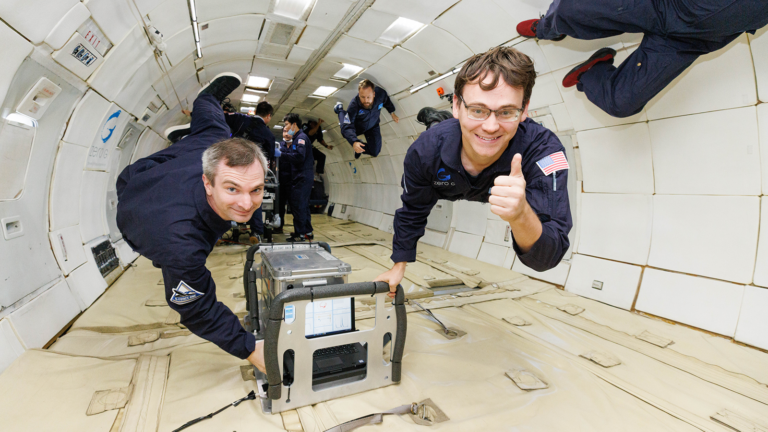 People in spacesuits float inside a zero-gravity aircraft, with one giving a thumbs-up and another interacting with equipment.