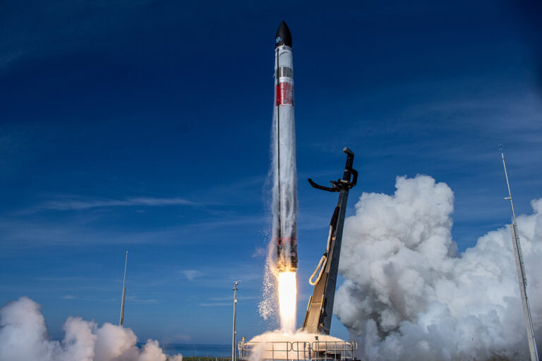 A rocket launches into the blue sky, emitting flames and smoke from its engines, with a support structure seen in the foreground.