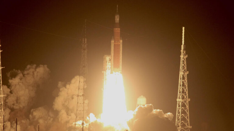 A spacecraft launches into the night sky, with bright flames and smoke emerging from its base, flanked by structures on either side.