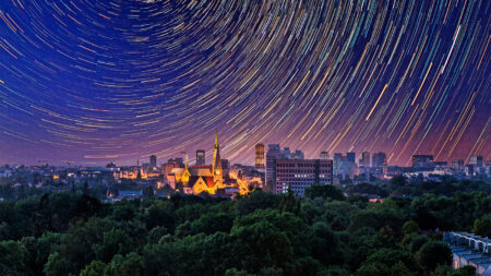 Night cityscape with star trails forming circles in the sky. A lit church and multiple buildings are visible amidst greenery.