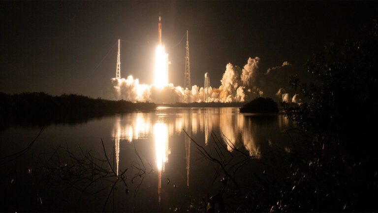 A rocket launches into the night sky, illuminating the surroundings and reflecting off a nearby body of water.