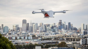 A white drone with four propellers flies above an urban area with a skyline of tall buildings in the background.