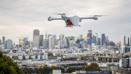 A white drone with four propellers flies above an urban area with a skyline of tall buildings in the background.