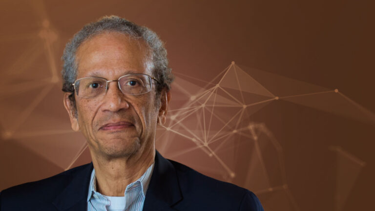 An older man with glasses and graying hair wears a dark suit and light blue shirt. He stands against a brown background with abstract geometric lines.