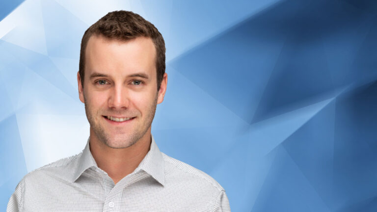 Man with short brown hair and a light beard is smiling at the camera, wearing a light gray dress shirt. The background is a blue geometric pattern.