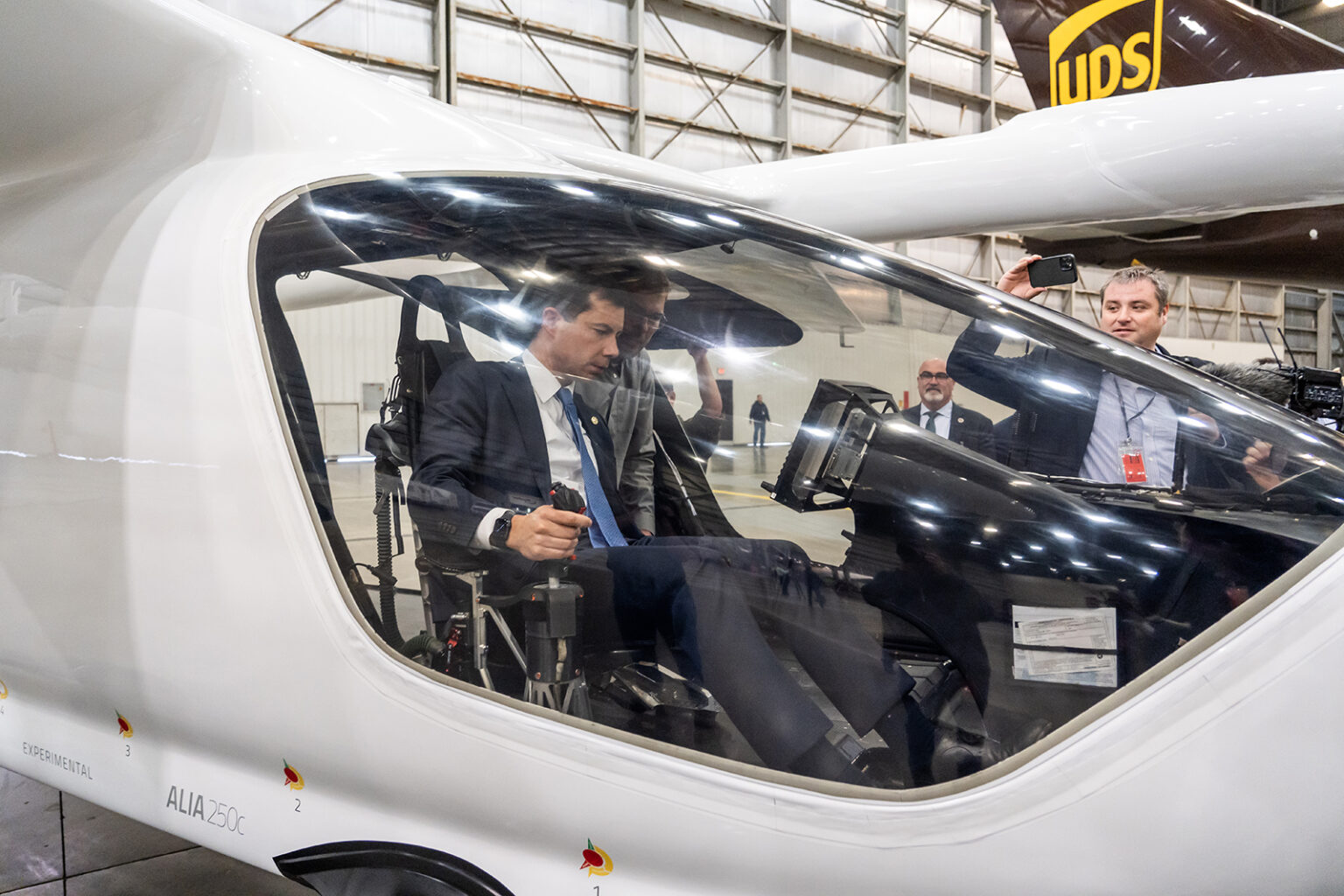 A man in a suit sits inside the cockpit of a sleek, white aircraft displayed in a large hangar, with onlookers and photographers nearby.