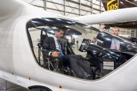 A man in a suit sits inside the cockpit of a sleek, white aircraft displayed in a large hangar, with onlookers and photographers nearby.