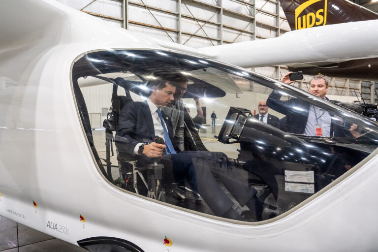 A man in a suit sits inside the cockpit of a sleek, white aircraft displayed in a large hangar, with onlookers and photographers nearby.