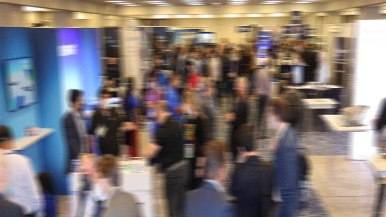 A busy trade show floor with people networking and visiting various exhibition booths in a large, well-lit venue.