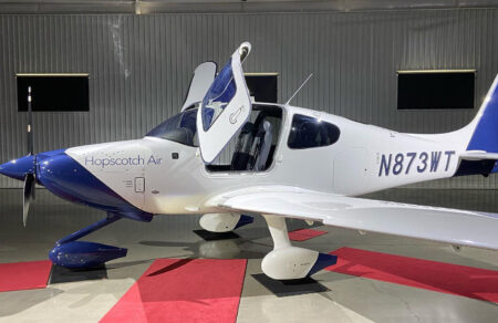 A small white and blue private aircraft with "Hopscotch Air" written on the side, parked in a hangar with open door and visible aircraft registration number N873WT.
