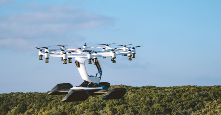 A multi-rotor drone, equipped with a seat and handlebars, hovers in the air against a backdrop of trees and a blue sky.