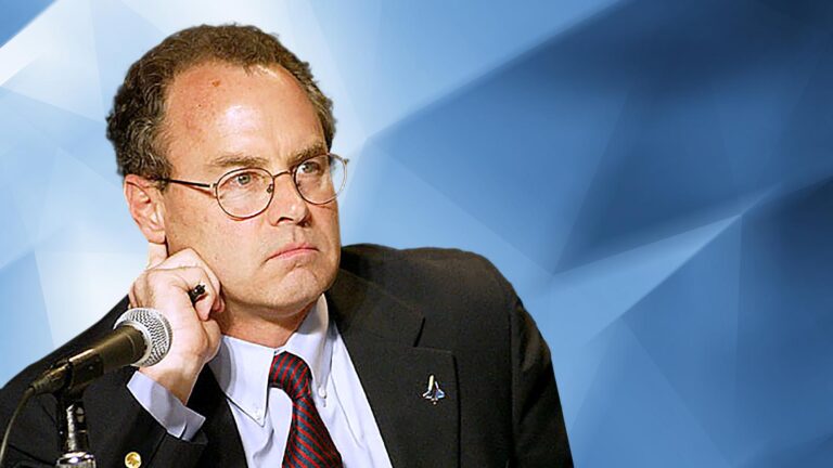 A man wearing glasses and a suit sits at a microphone with his hand resting on his cheek against a geometric blue background.