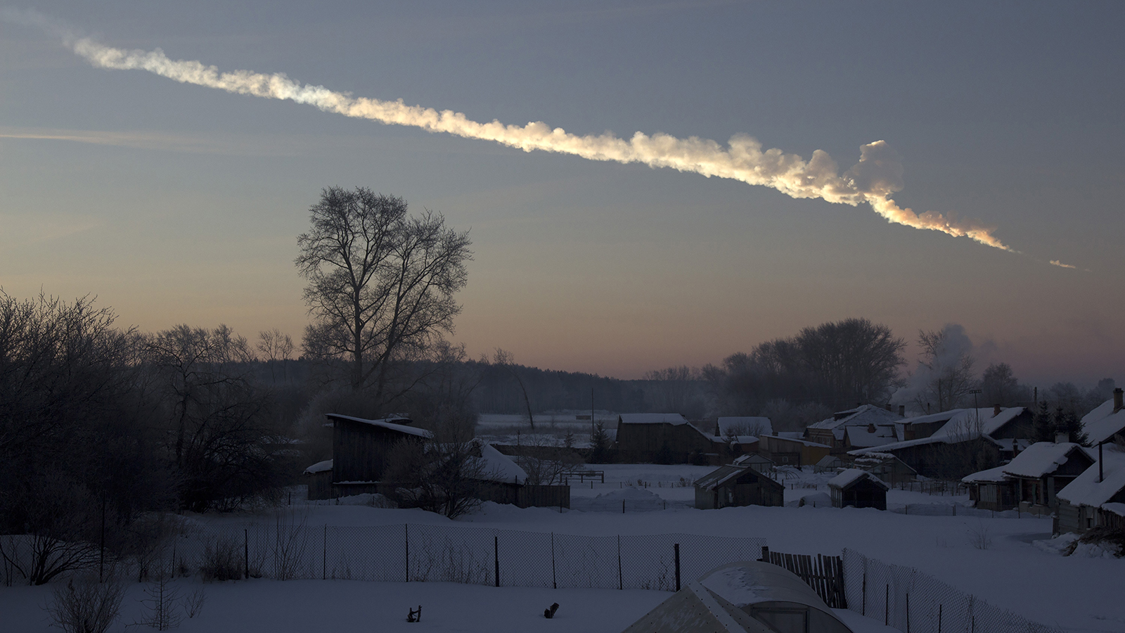 A bright streak of smoke stretches across the dawn sky, over a quiet snow-covered village with trees and scattered houses.