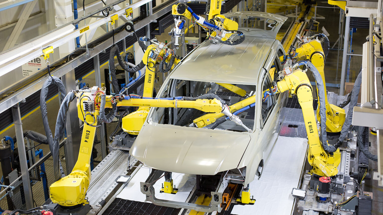 Robotic arms work on an unfinished car frame on an assembly line in a factory setting.
