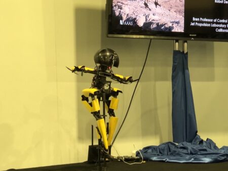 A yellow and black humanoid robot stands on a stage near a monitor displaying information about a professor and the Jet Propulsion Laboratory.