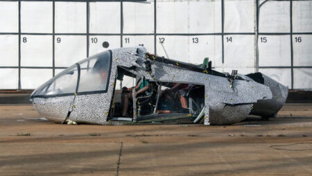 A damaged aircraft fuselage lies on the ground in front of a numbered wall grid, showing signs of a severe impact or crash test.