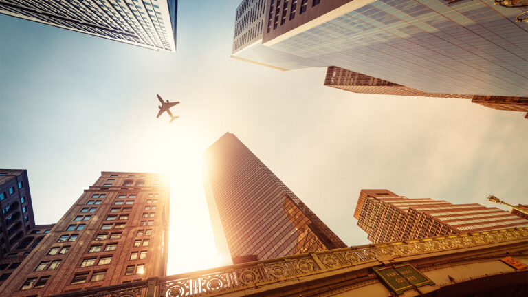 A plane flies overhead amid tall skyscrapers under a bright, sunny sky; the viewpoint is from street level looking up.