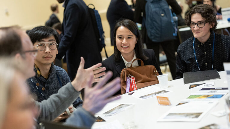 A group of people sit around a table engaged in conversation, some with notebooks and pamphlets in front of them, in a busy indoor setting.