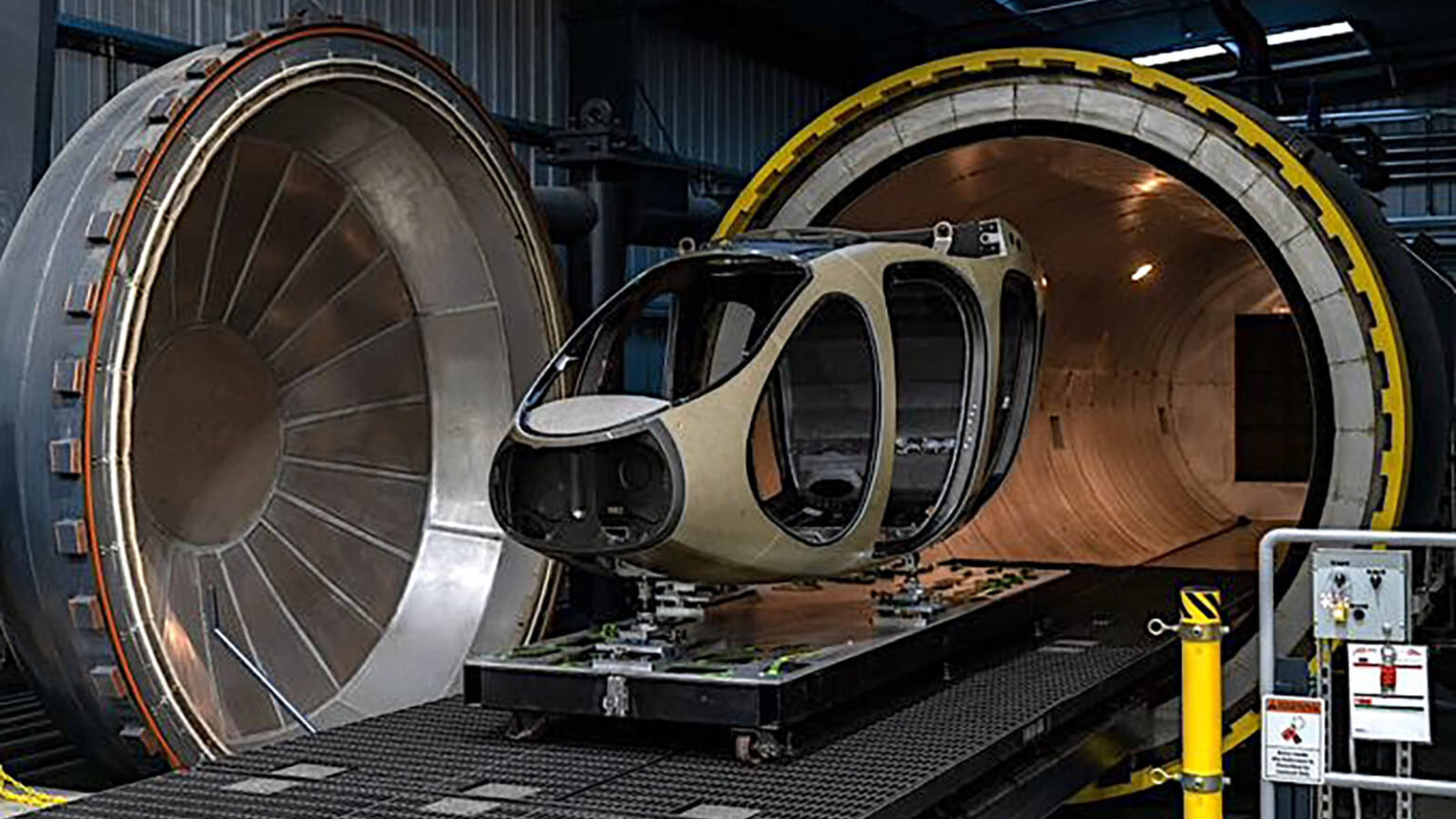 A small, futuristic aircraft fuselage is positioned between two large, cylindrical industrial machines in a manufacturing facility.