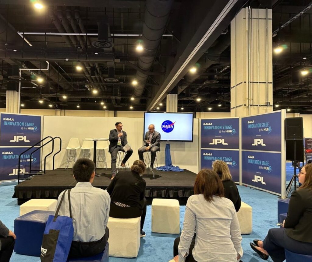 Two speakers are seated on a stage during a NASA JPL event, speaking in front of an audience in an indoor conference setting.