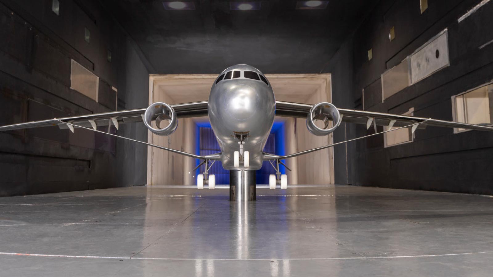 Front view of an aircraft model in a wind tunnel testing environment. The model is mounted on a support and surrounded by dark walls, with blue lighting in the background.