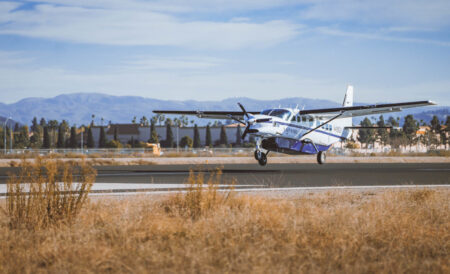 A small propeller airplane takes off from a runway in a dry, grassy area with mountains in the background.