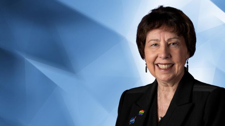 A woman with short dark hair wearing a black blazer smiles at the camera in front of a geometric blue background.