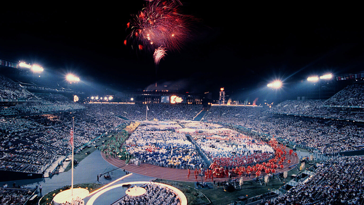 A nighttime stadium scene with athletes in colorful uniforms on the field, fireworks overhead, and the stands filled with spectators.