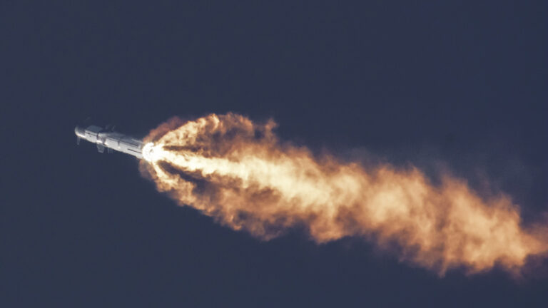 A rocket is in flight with its engines producing a bright flame and trail of exhaust smoke against a dark sky.