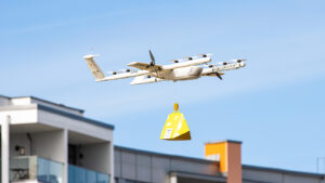 A drone flying in the sky, carrying a yellow package. It is near a building with balconies visible. The sky is clear with no clouds.