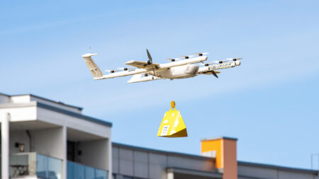 A drone flying in the sky, carrying a yellow package. It is near a building with balconies visible. The sky is clear with no clouds.