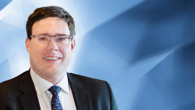 A man wearing glasses, a suit, and a tie is smiling against a geometric blue background.