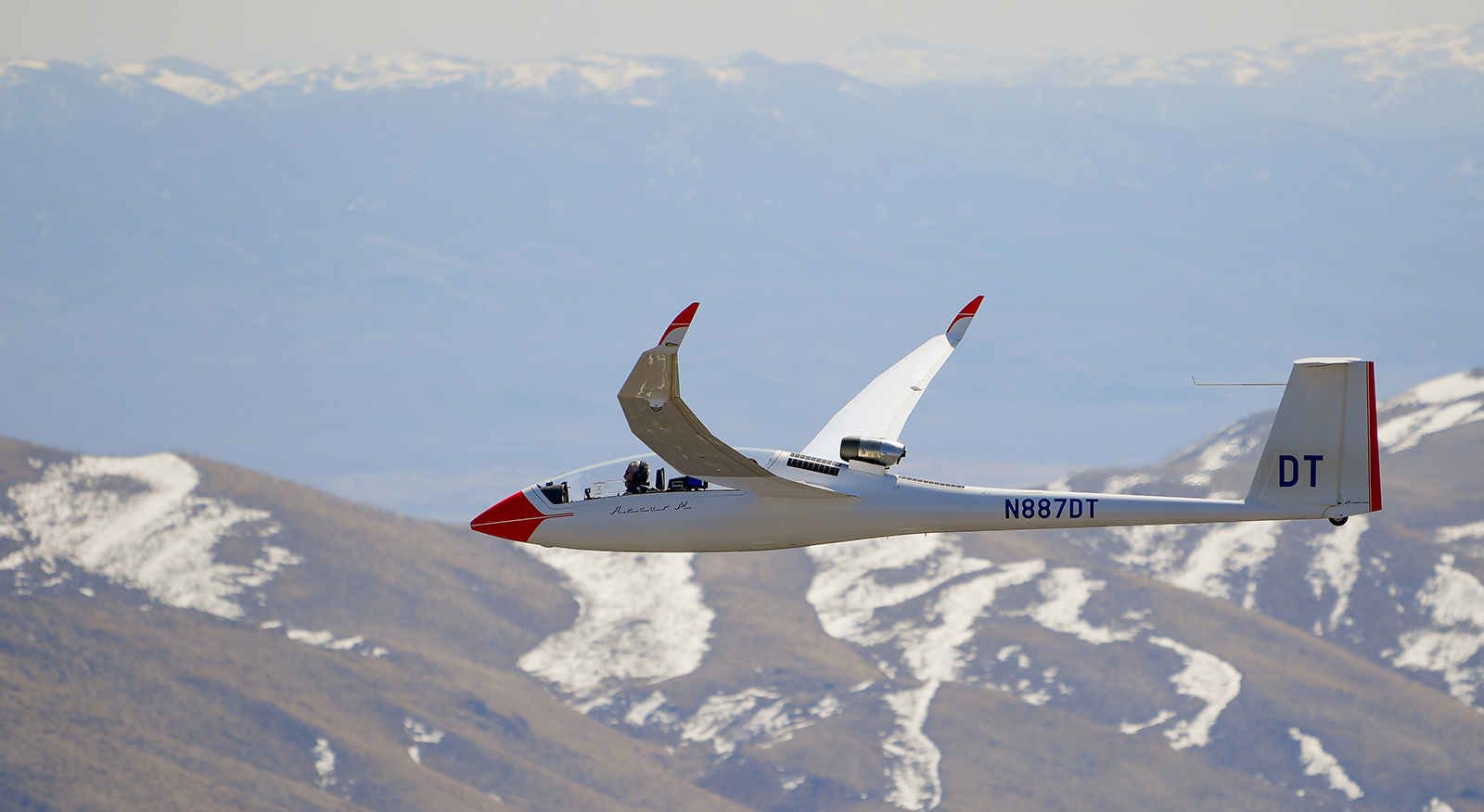 A glider with the registration N887DT flying over a mountainous landscape partially covered in snow.