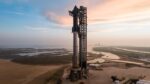 A large rocket stands upright on its launch pad with a tall supporting structure beside it, set against a backdrop of a coastal landscape at sunset.