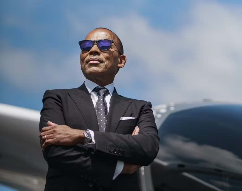A man in a black suit and polka-dot tie stands confidently with folded arms in front of an airplane, wearing sunglasses on a clear day.