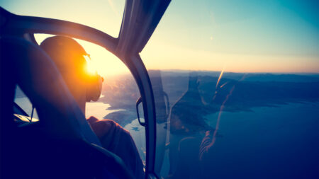 A person is seated in a helicopter, wearing headphones while flying over a coastal landscape with mountains and water bodies at sunset.