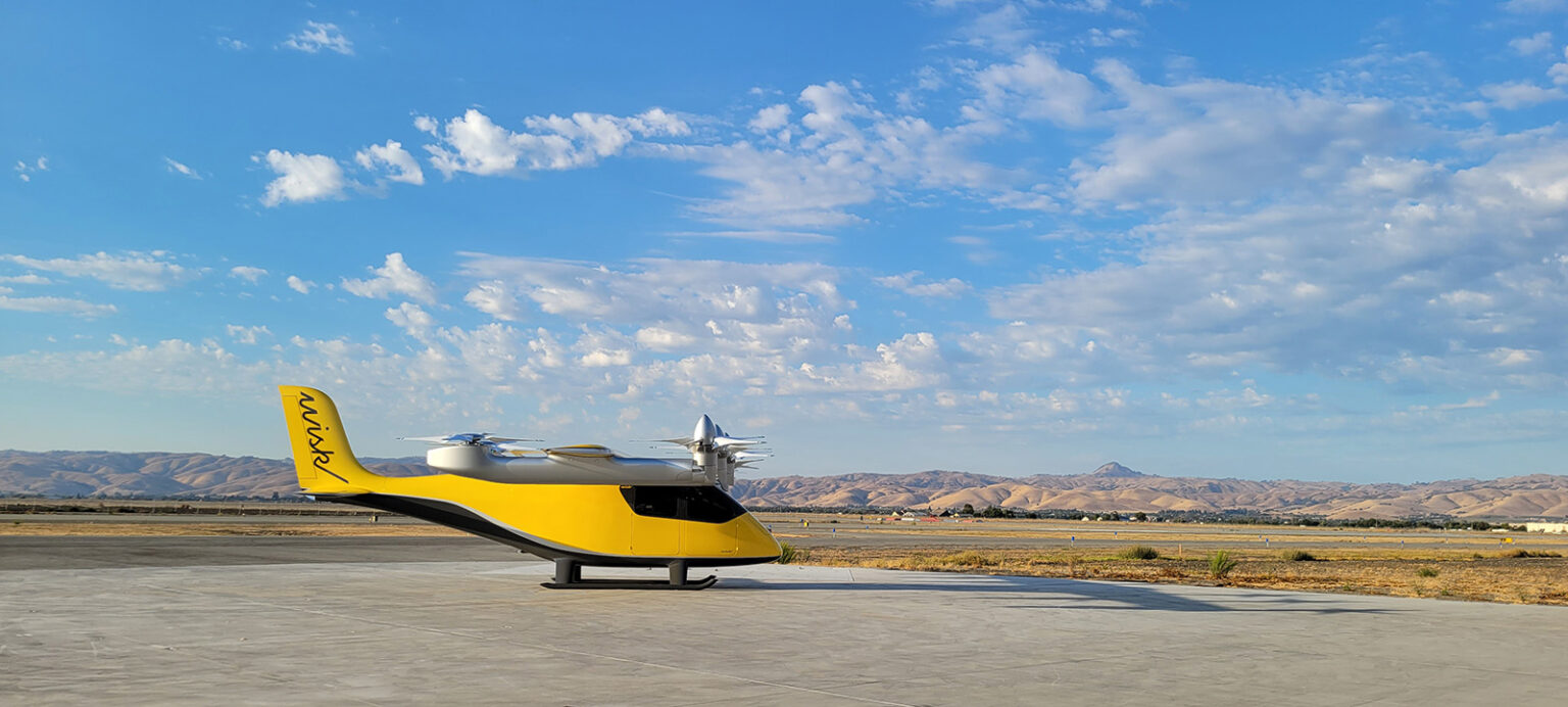 A yellow, electric vertical takeoff and landing (eVTOL) aircraft is parked on an expansive runway with a clear, blue sky and mountainous landscape in the background.