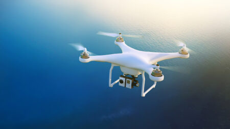 A white quadcopter drone with a camera flies over a body of water under a clear sky.