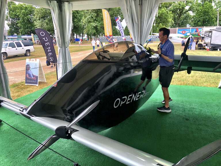 A man in a blue shirt inspects a sleek, black Opener aircraft displayed under a white tent with other exhibits and vehicles visible in the background.